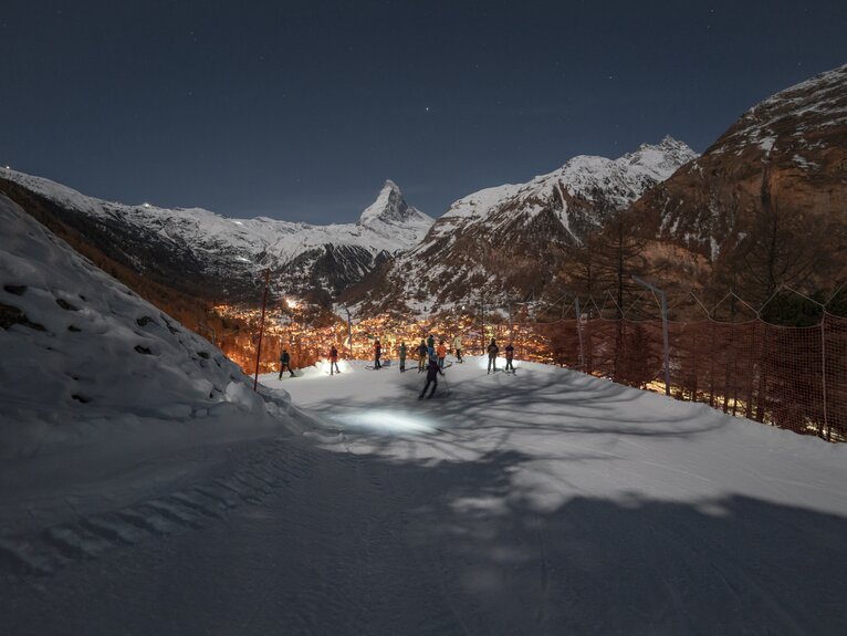 Piste dans l'obscurité avec des skieurs regardant vers le bas Zermatt illuminé, le Cervin en arrière-plan | © Gabriel_Perren