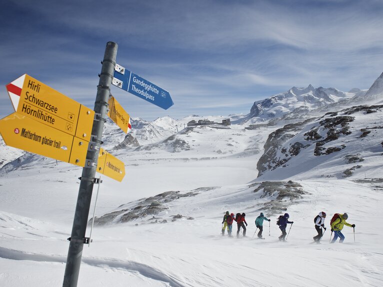 Gruppe von Schneeschuhläufer kämpft sich durch den Schnee, im Vordergrund steht ein Wegweiser | © Michi_Portmann